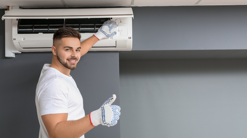 A male technician wearing a white shirt and gloves stands next to the interior part of a split air conditioner and holds his thumb up.